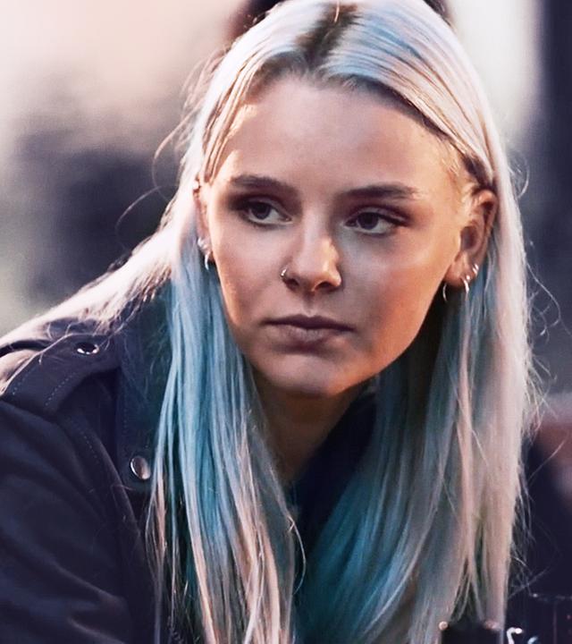 Eine junge Frau mit grau-blond gefärbten Haaren (Olivia Burkhart) spricht mit einem jungen Mann (Louis Held).