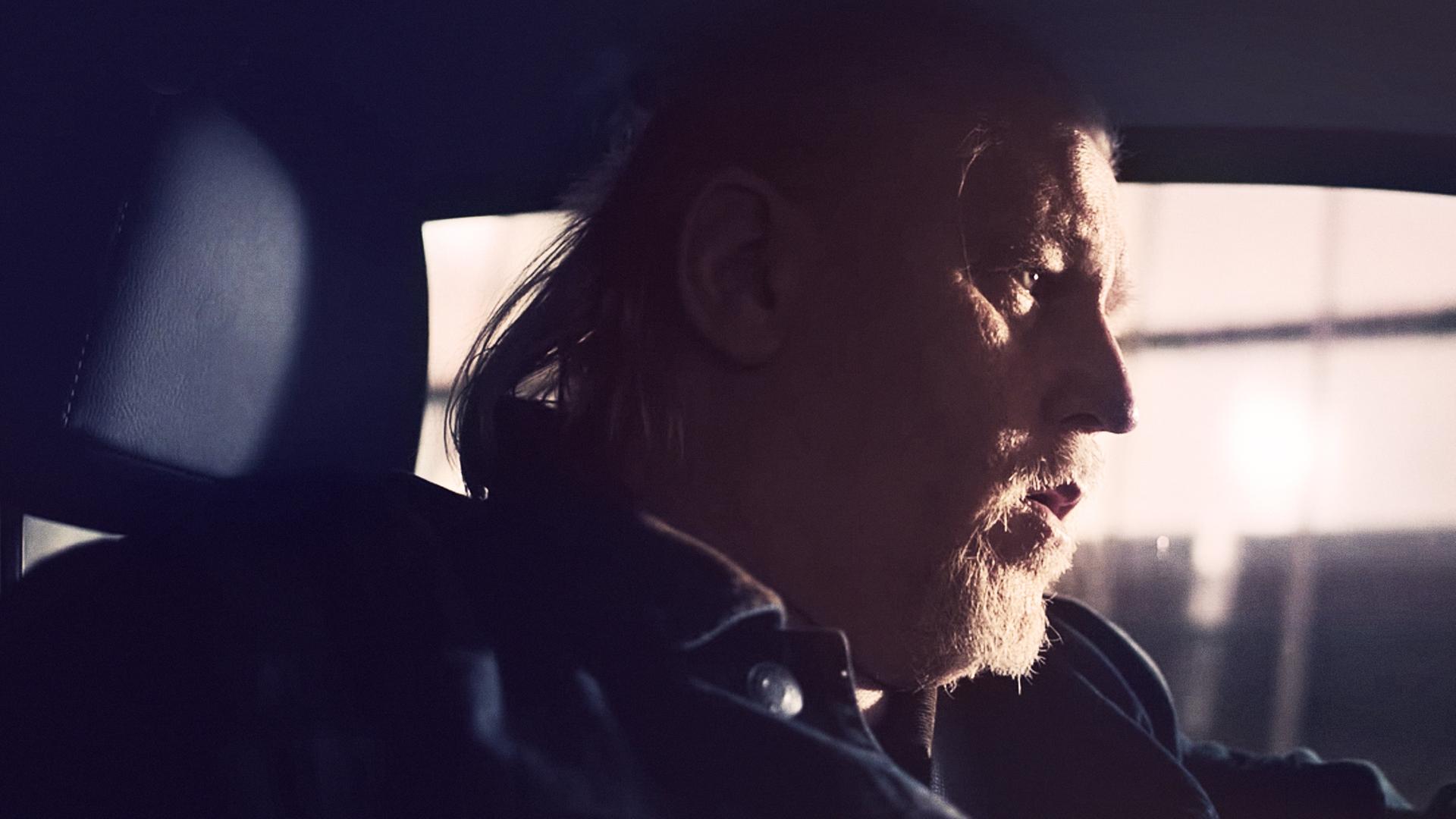 Ein Mann mit weißem Bart und längeren weißen Haaren (Arved Birnbaum) sitzt auf dem Fahrersitz eines Autos. Man sieht ihn aus dem Profil.