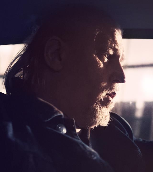 Ein Mann mit weißem Bart und längeren weißen Haaren (Arved Birnbaum) sitzt auf dem Fahrersitz eines Autos. Man sieht ihn aus dem Profil.