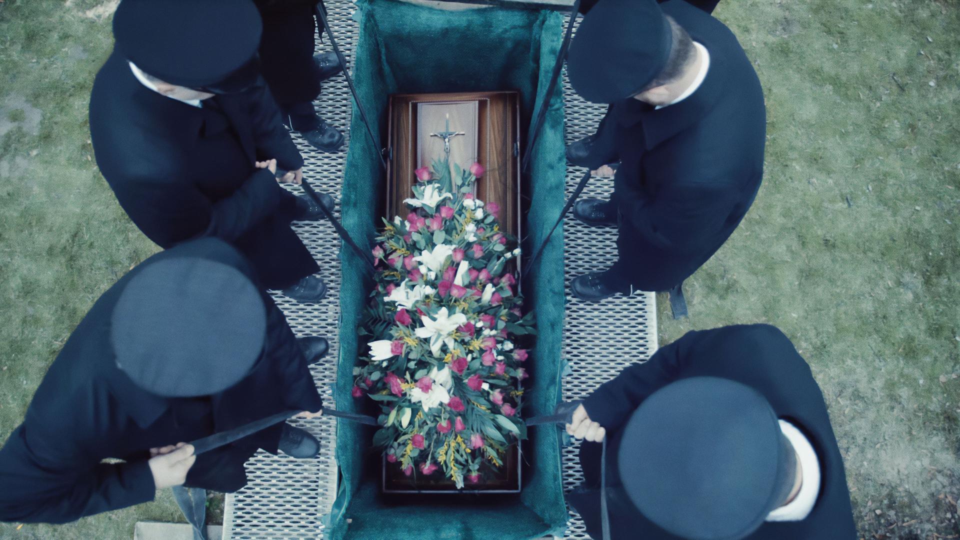 Aufsicht auf ein geöffnetes Grab, in das gerade ein Sarg mit buntem Blumenschmuck heruntergelassen wird.