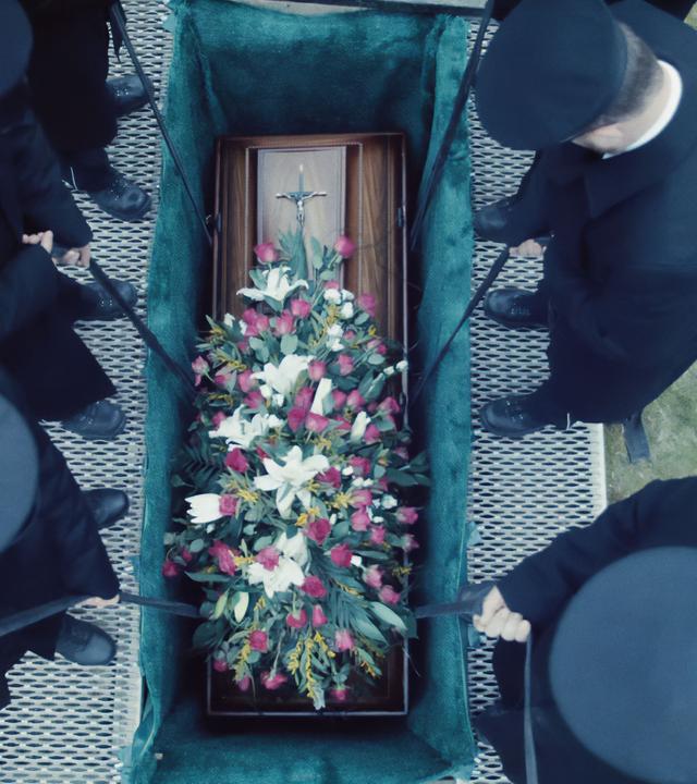 Aufsicht auf ein geöffnetes Grab, in das gerade ein Sarg mit buntem Blumenschmuck heruntergelassen wird.