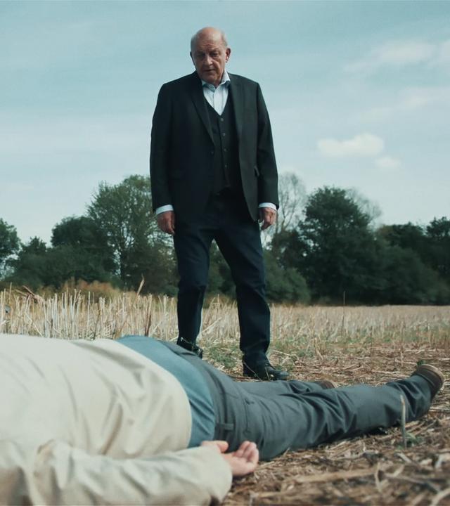 Wilsberg (Leonard Lansink) und Tilker (Patricia Meeden) stehen in einem Stoppelfeld. Vor ihnen liegt bäuchlinks die Leiche eines Mannes. 