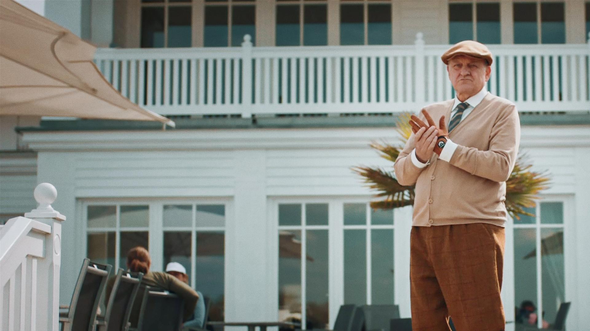 Wilsberg (Leonard Lansink) in einem altmodischen Golfer-Outfit auf einer Terrasse. Stühle und Tische, Sonnenschirme und ein weißes Haus im Hintergrund.