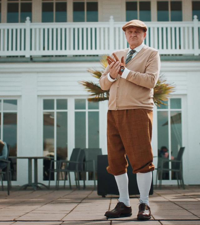 Wilsberg (Leonard Lansink) in einem altmodischen Golfer-Outfit auf einer Terrasse. Stühle und Tische, Sonnenschirme und ein weißes Haus im Hintergrund.