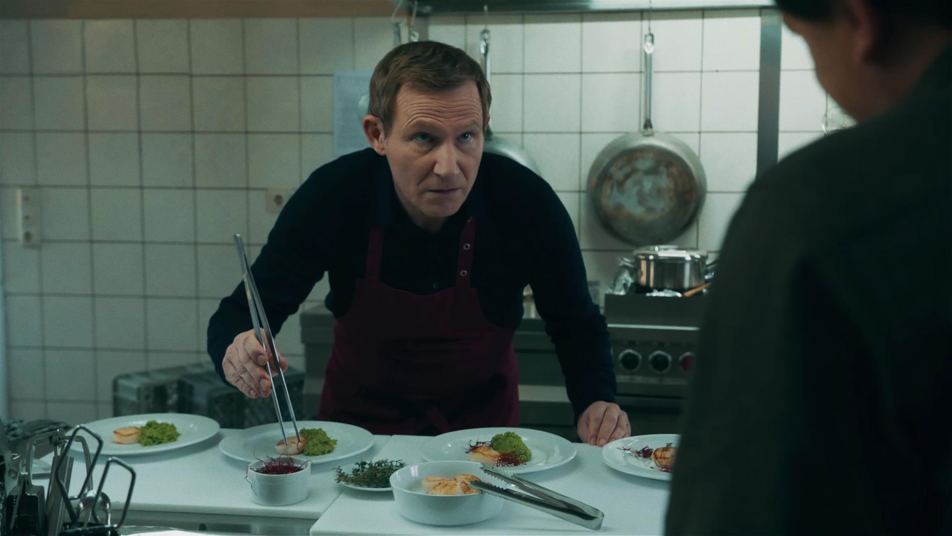 In einer Restaurantküche richtet ein schwarz-gekleidet Koch das Essen auf vier Tellern an. Dabei scheint er von einer unbekannten Person am rechten Bildrand erwischt zu werden. 