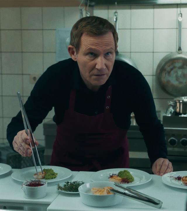 In einer Restaurantküche richtet ein schwarz-gekleidet Koch das Essen auf vier Tellern an. Dabei scheint er von einer unbekannten Person am rechten Bildrand erwischt zu werden. 