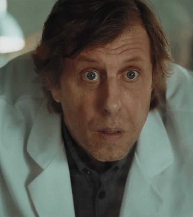 Ekki (Oliver Korittke) in einem weißen Laborkittel, sein Gesichtsausdruck erstaunt und geschockt. 