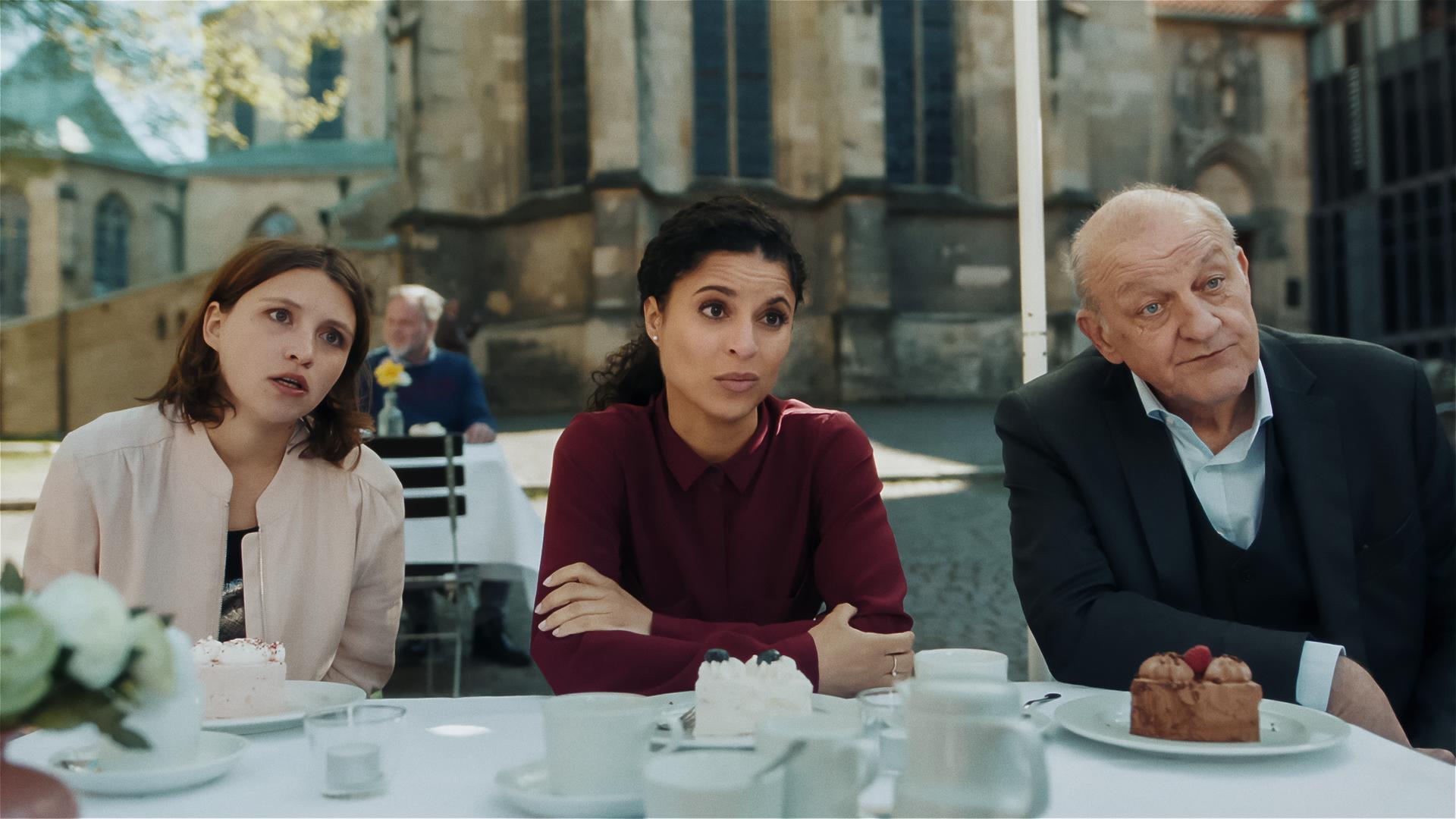 Merle (Janina Fautz), die eine junge Frau mit braunen Haaren ist, Tessa Tilker (Patricia Meeden) und Wilsberg (Leonard Lansink) sitzen in einem Café auf dem Marktplatz, Tortenstücke auf dem Tisch vor ihnen, und schauen zu etwas außerhalb des Bildes. Alle haben ungläubige und überraschte Gesichtsausdrücke.