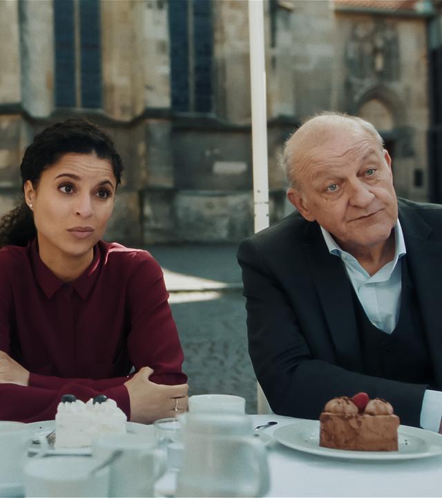 Merle (Janina Fautz), die eine junge Frau mit braunen Haaren ist, Tessa Tilker (Patricia Meeden) und Wilsberg (Leonard Lansink) sitzen in einem Café auf dem Marktplatz, Tortenstücke auf dem Tisch vor ihnen, und schauen zu etwas außerhalb des Bildes. Alle haben ungläubige und überraschte Gesichtsausdrücke.