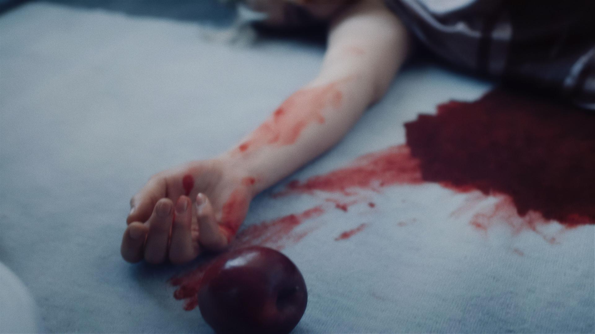 Auf einer blutgetränkten Matratze liegt eine Frau, von der man nur den ausgestreckten, blutbeschmierten Arm sehen kann. Vor der Hand liegt ein roter Apfel.