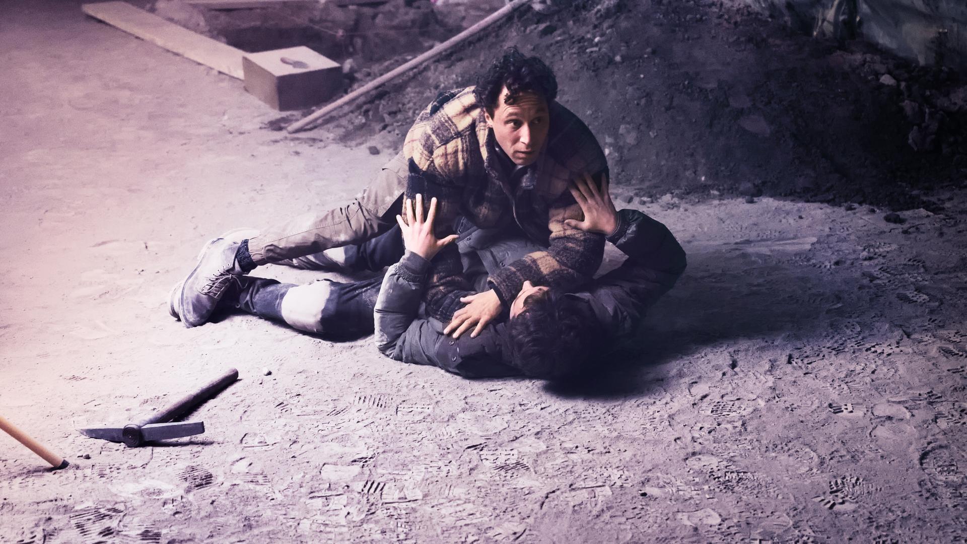 Auf dem sandigen Boden einer Baustelle drückt ein Mann einen anderen nieder.