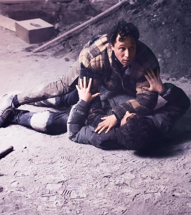 Auf dem sandigen Boden einer Baustelle drückt ein Mann einen anderen nieder.