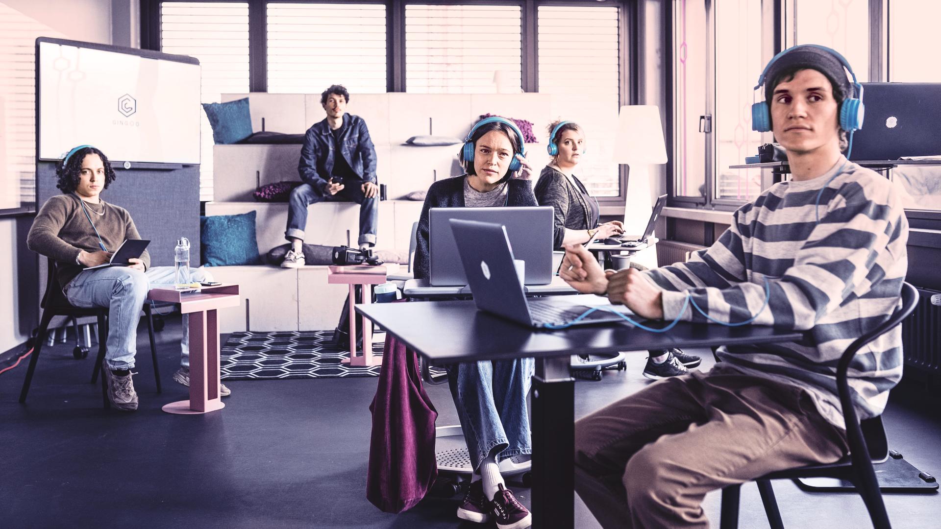 In einem Coworking-Space mit Tischen und Sofas sitzen fünf junge Menschen mit Laptops und blauen Kopfhörern, die irritiert zu etwas außerhalb des Bildes schauen.