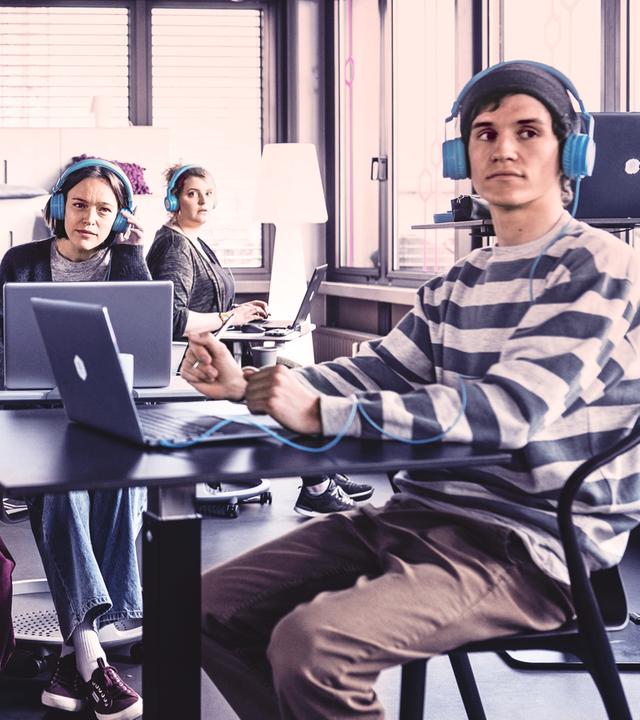In einem Coworking-Space mit Tischen und Sofas sitzen fünf junge Menschen mit Laptops und blauen Kopfhörern, die irritiert zu etwas außerhalb des Bildes schauen.