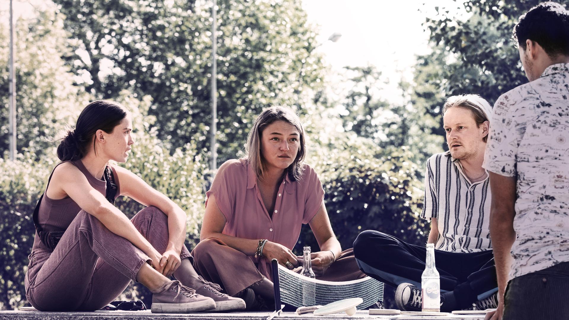 Im Park sitzt auf dem Boden eine Gruppe junger Menschen: zwei Frauen links und zwei Männer rechts. Es ist Sommer.