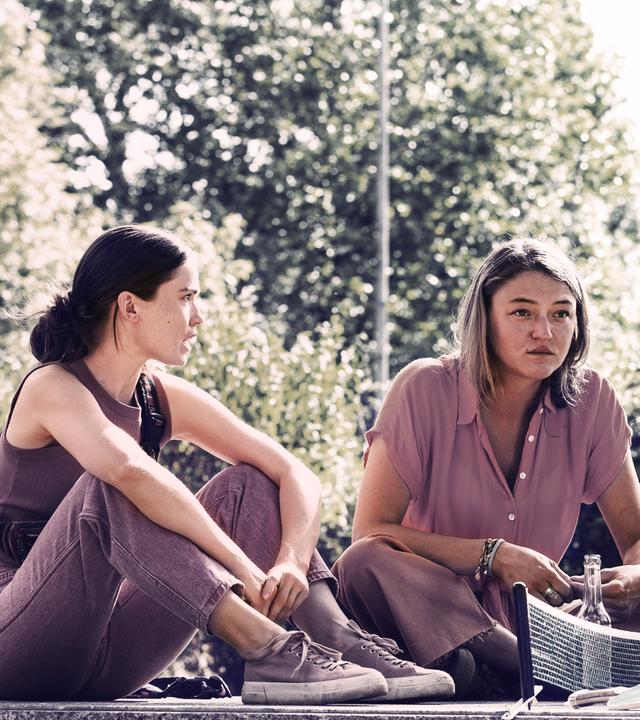 Im Park sitzt auf dem Boden eine Gruppe junger Menschen: zwei Frauen links und zwei Männer rechts. Es ist Sommer.