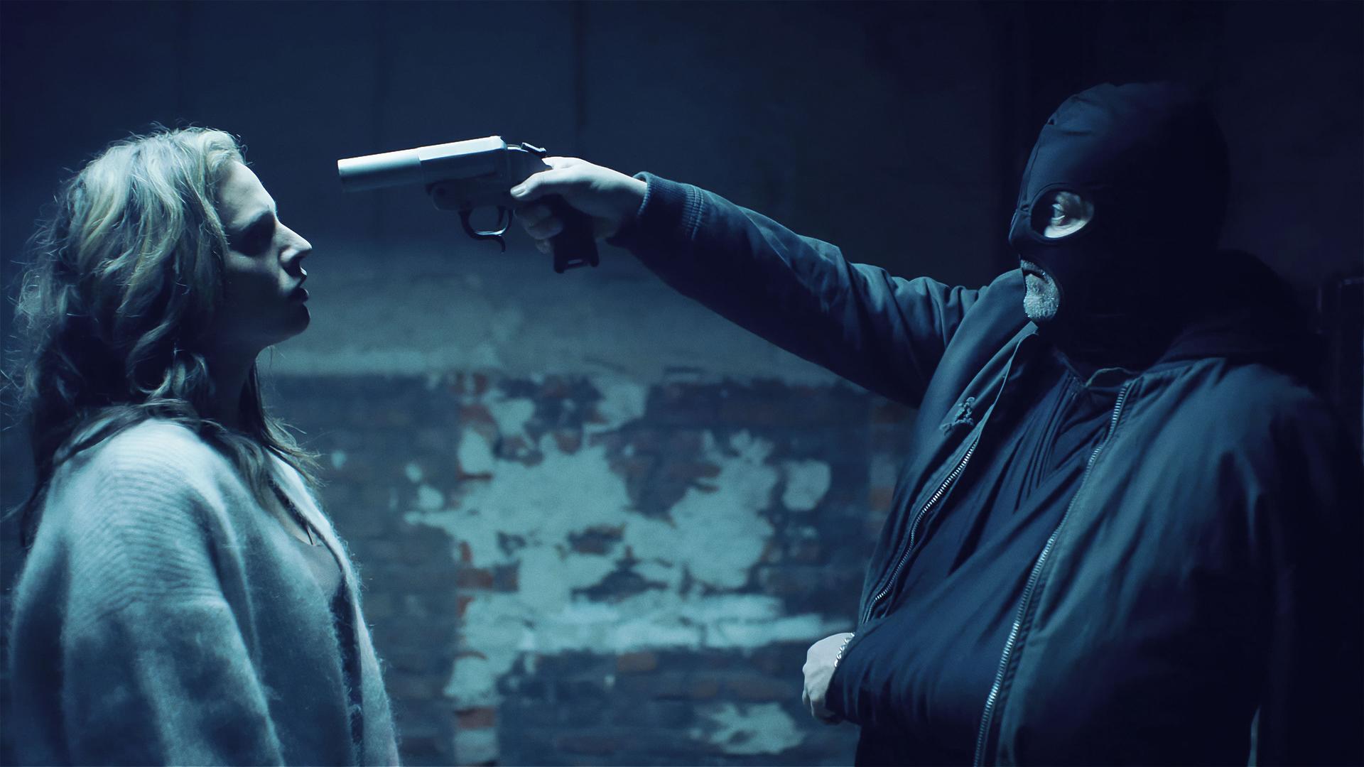 Eine Frau und ein maskierter Mann stehen sich in einem heruntergekommenen Raum gegenüber. Die Frau trägt eine Stoffjacke; der Mann trägt neben der schwarzen Maske eine Bomberjacke. Sein linker Arm ist in eine Bandage gelegt und mit der anderen Hand hält er einen Revolver, den er auf die Frau gerichtet hat.