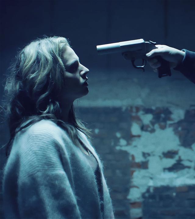 Eine Frau und ein maskierter Mann stehen sich in einem heruntergekommenen Raum gegenüber. Die Frau trägt eine Stoffjacke; der Mann trägt neben der schwarzen Maske eine Bomberjacke. Sein linker Arm ist in eine Bandage gelegt und mit der anderen Hand hält er einen Revolver, den er auf die Frau gerichtet hat.