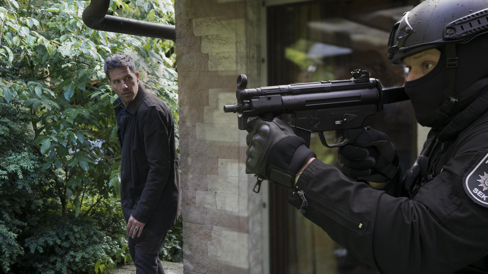 Oliver von Bodenstein (Tim Bergmann) steht hinter einer Hausmauer und schaut von dieser auf einen Polizisten vom SEK in Einsatzkleidung. Dieser hält ein automatisches Maschinengewehr in Schussposition.