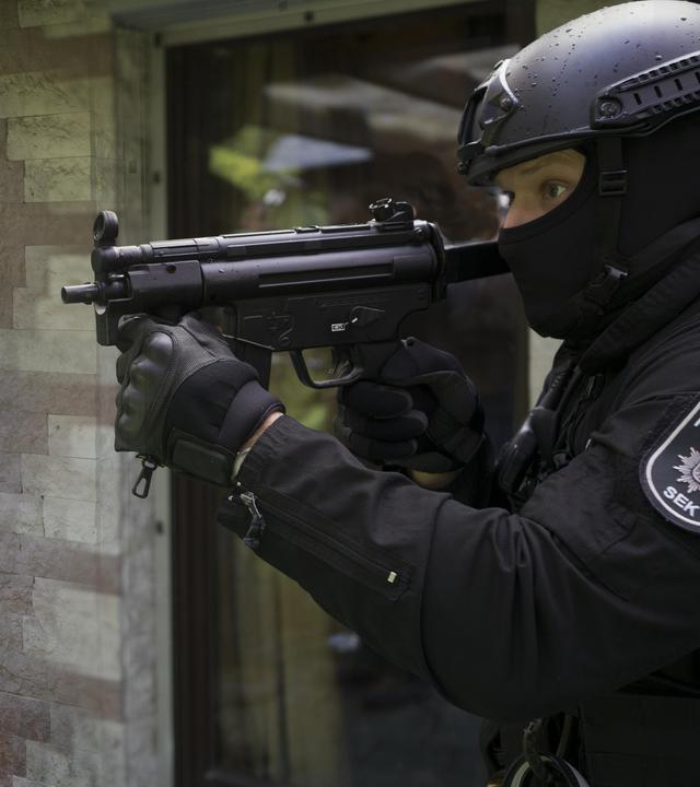 Oliver von Bodenstein (Tim Bergmann) steht hinter einer Hausmauer und schaut von dieser auf einen Polizisten vom SEK in Einsatzkleidung. Dieser hält ein automatisches Maschinengewehr in Schussposition.