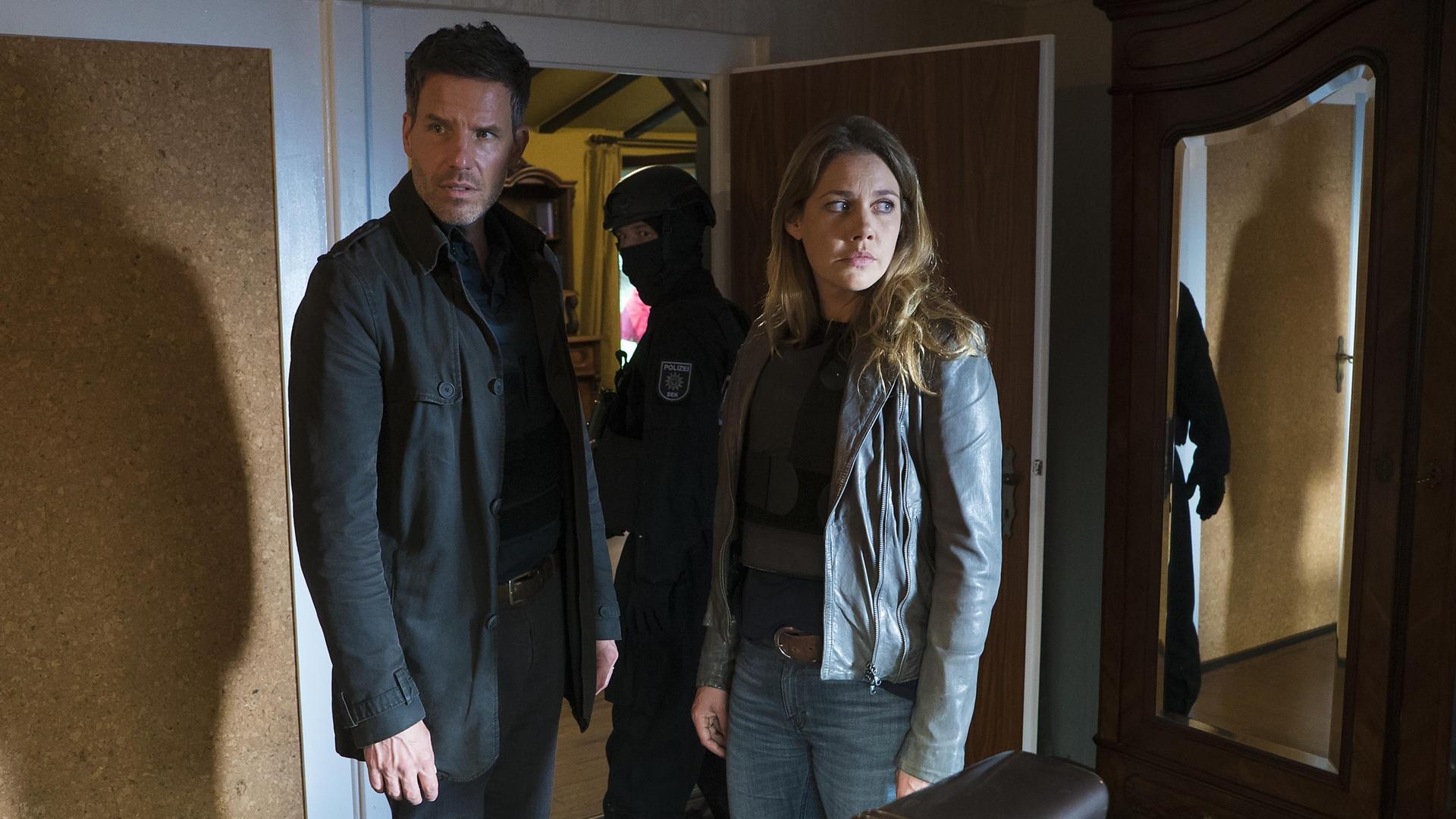 Oliver von Bodenstein (Tim Bergmann) und Pia Kirchhoff (Felicitas Woll) stehen in einem Zimmer und schauen sich überrascht um. Hinter ihnen ist ein Polizist in der offenen Tür zu sehen.