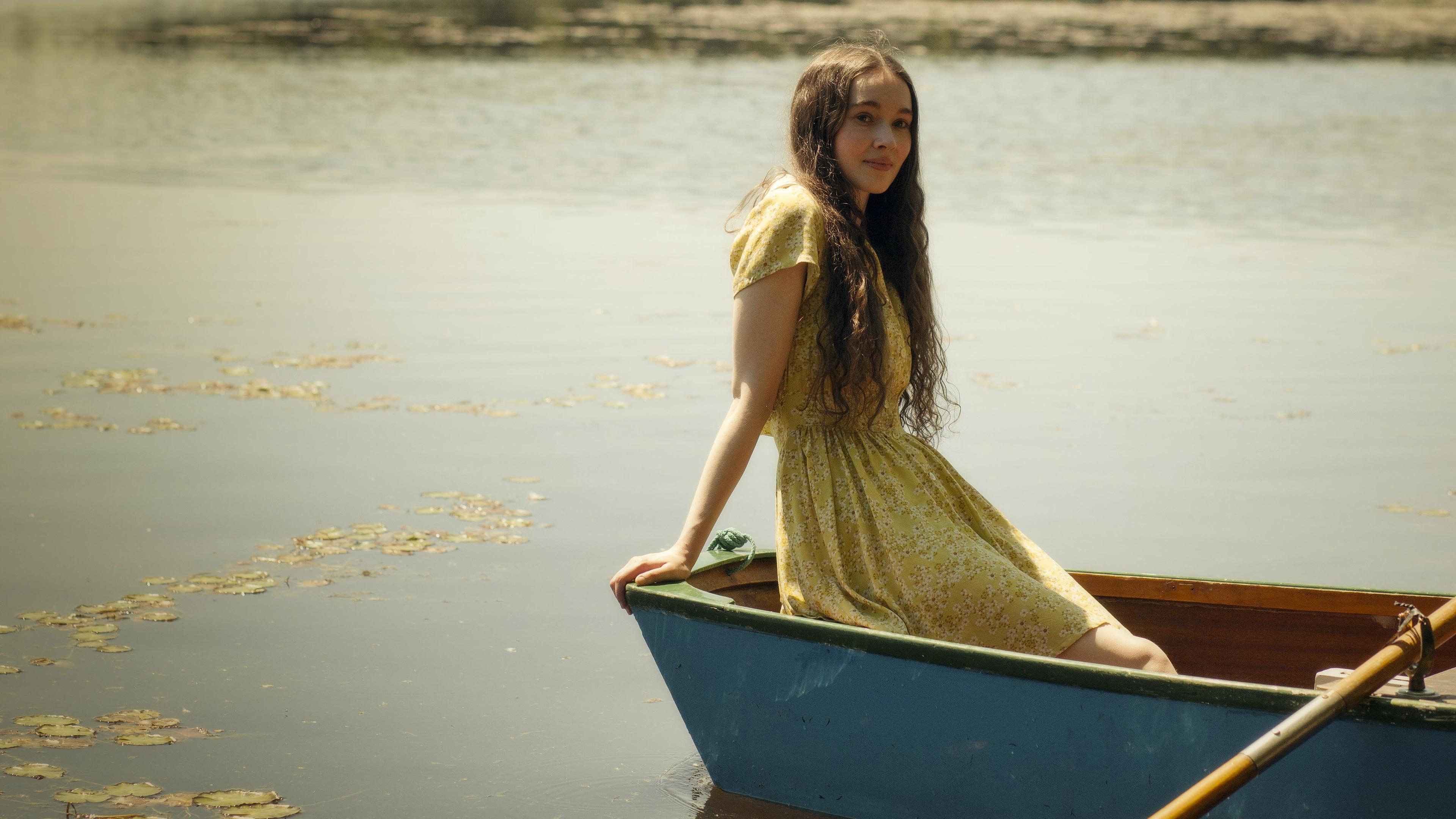 Nora (Julia Anna Grob) sitzt auf ihren Armen abgestützt in einem Sommerkleid in einem Kanu, das auf dem Wasser eines Sees treibt. 