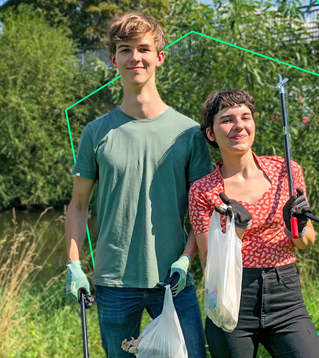 Maximo (l.) und Ella (r.) stehen auf einer Wiese am Ufer eines Flusses. Sie halten gefüllte Müllbeutel und Greifzangen in der Hand und lächeln in die Kamera.