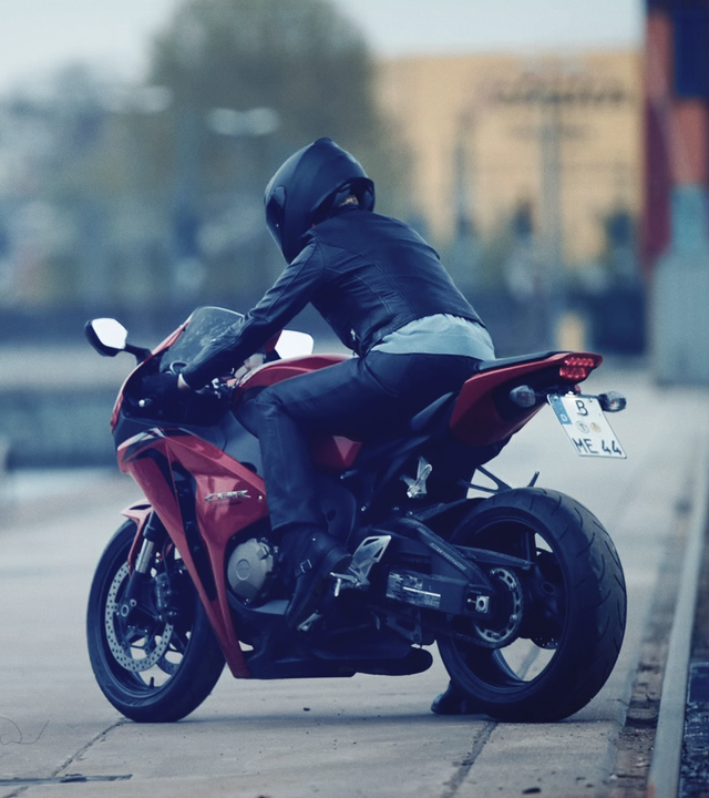 An einem Flussufer steht ein rotes Motorrad mit einem schwarzgekleideten Fahrer.