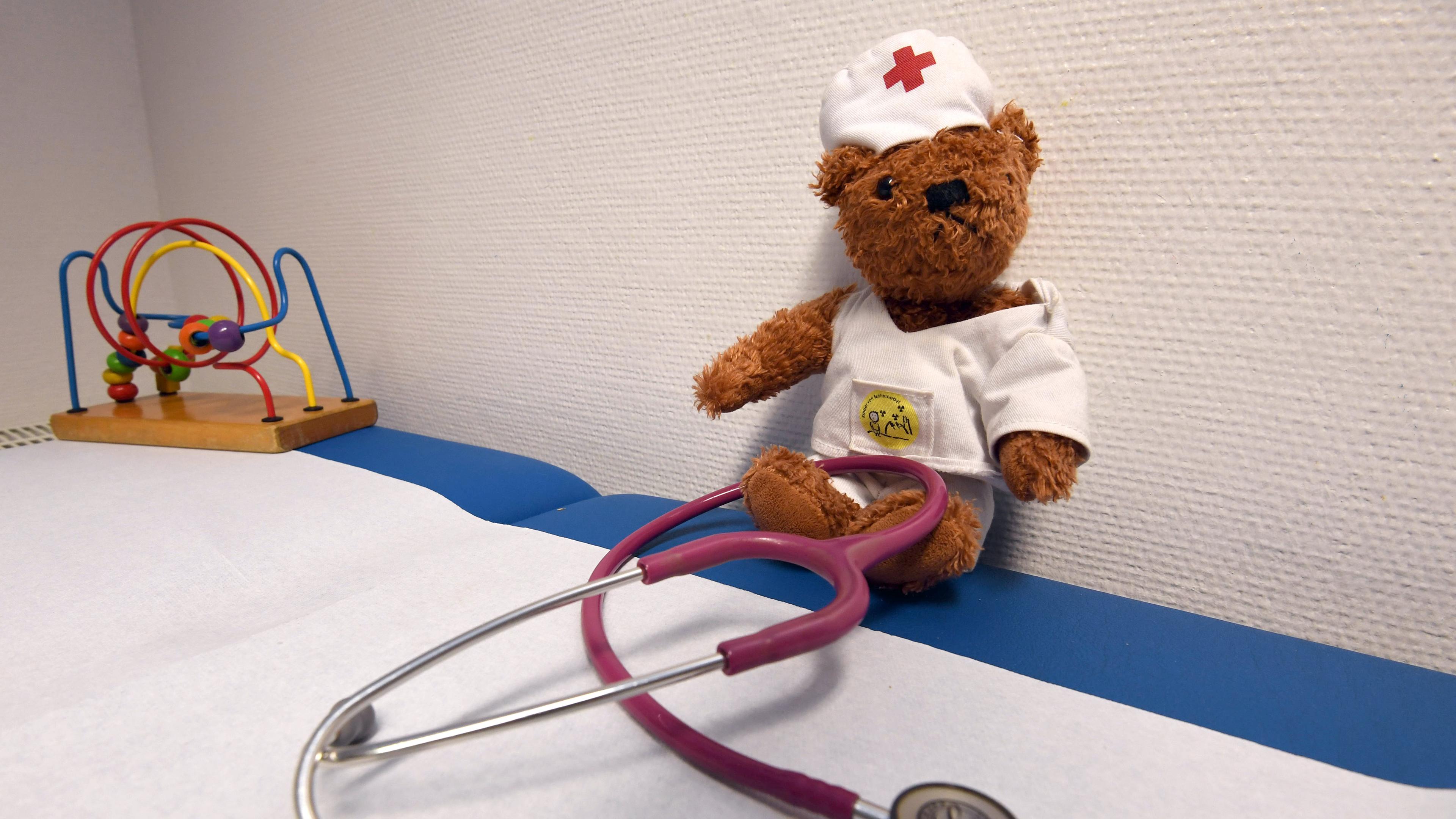 Ein Stethoskop liegt in der Praxis einer Kinderärztin vor einem als Arzt verkleideten Teddy auf einer Liege.