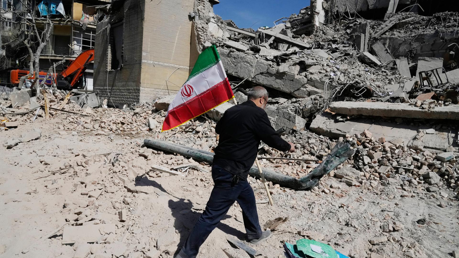 Ein Mann läuft durch den Schutt eines zerstörten Hauses und trägt die grün-weiß-rote Flagge Irans.