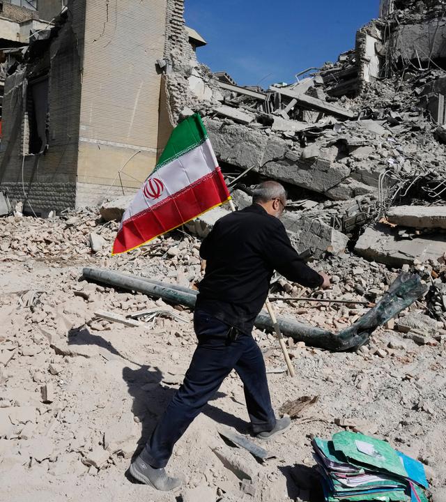 Ein Mann läuft durch den Schutt eines zerstörten Hauses und trägt die grün-weiß-rote Flagge Irans.