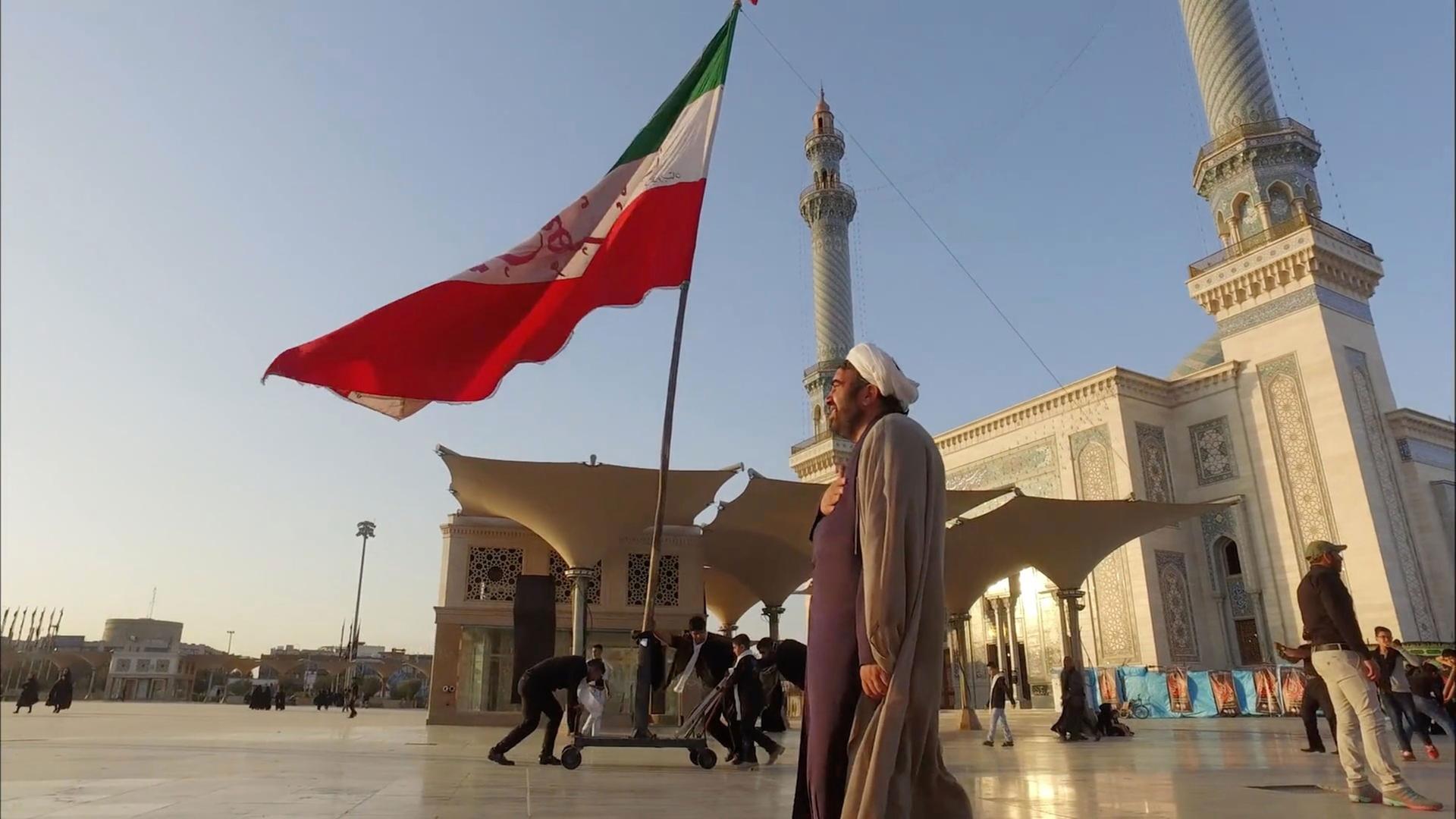 "Teheran extrem - Subkultur im Gottesstaat": Ein Mullah steht auf einem Platz vor einem Tempel und einer iranischen Flagge.