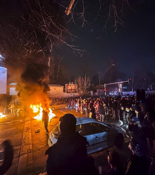 Auf einem Platz bei Nacht haben sich Menschen versammelt, auf einem Parkplatz brennen mehrere Feuer.
