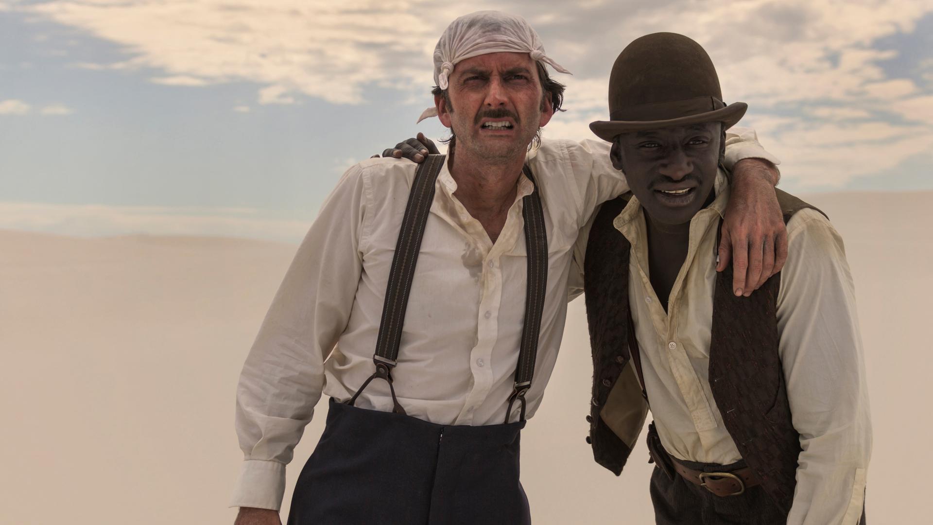 Phileas Fogg und sein Diener Jean Passepartout taumeln durstig durch die Wüste und haben sich gegenseitig stützend die Arme um die Schultern gelegt.