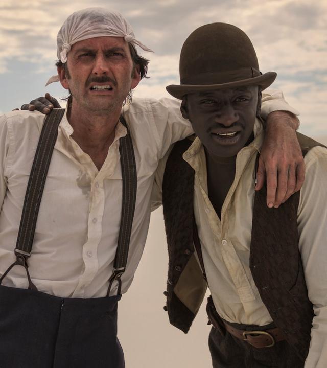 Phileas Fogg und sein Diener Jean Passepartout taumeln durstig durch die Wüste und haben sich gegenseitig stützend die Arme um die Schultern gelegt.