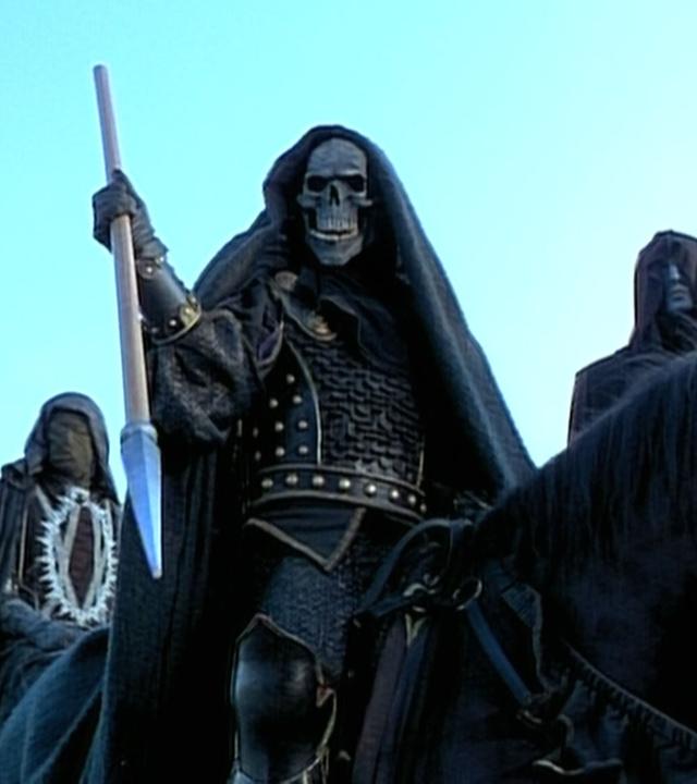 Drei Reiter auf schwarzen Pferden mit schwarzen Mänteln und Masken. Der Reitter in der Mitte trägt eine Totenkopfmaske und hält einen Speer in der Hand.