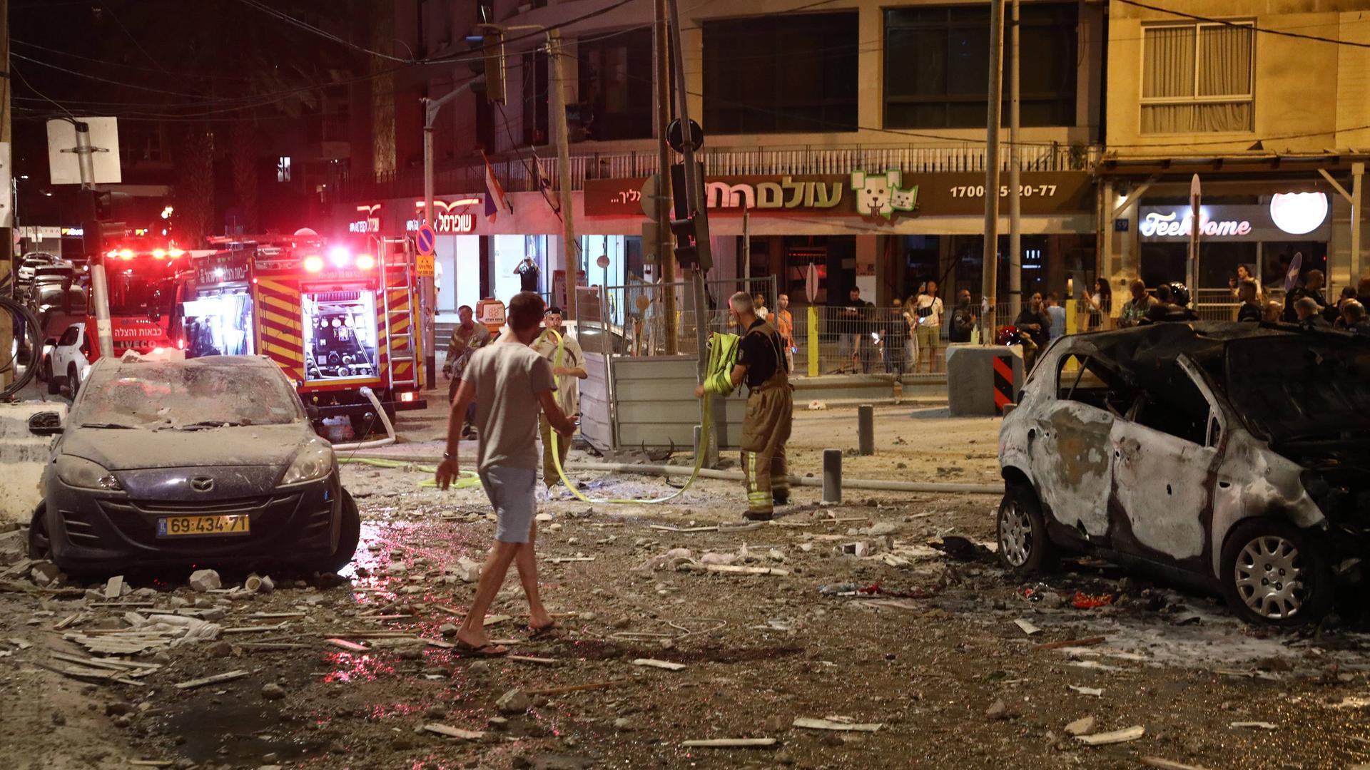 Eine Straßenkreuzung in Tel Aviv bei Nacht, nach einem Raketenangriff: Am Straßenrand stehen beschädigte Autos, Menschen begutachten die Schäden an Gebäuden und Straße.