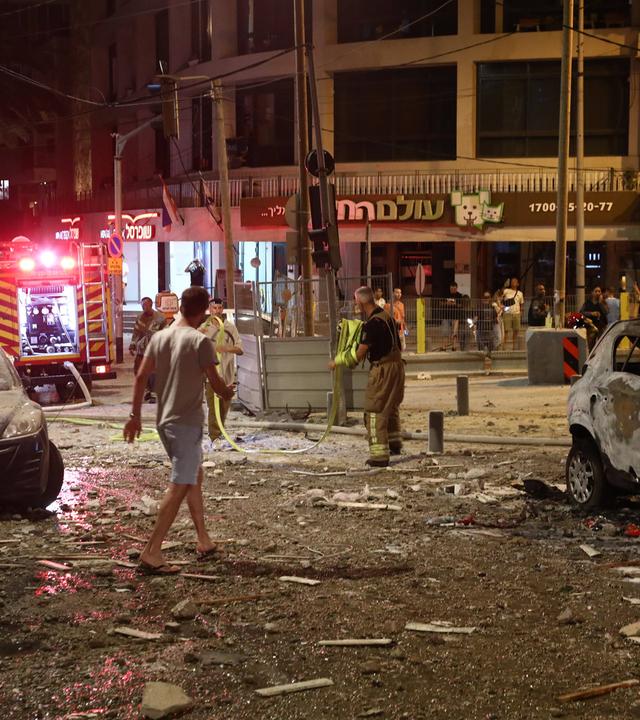 Eine Straßenkreuzung in Tel Aviv bei Nacht, nach einem Raketenangriff: Am Straßenrand stehen beschädigte Autos, Menschen begutachten die Schäden an Gebäuden und Straße.