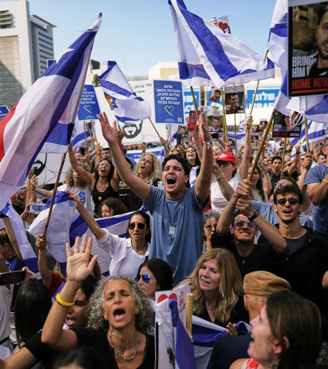 Auf einem Platz hat sich eine Menschenmenge versammelt, die weiß-blaue Fahnen schwenkt und jubelt.
