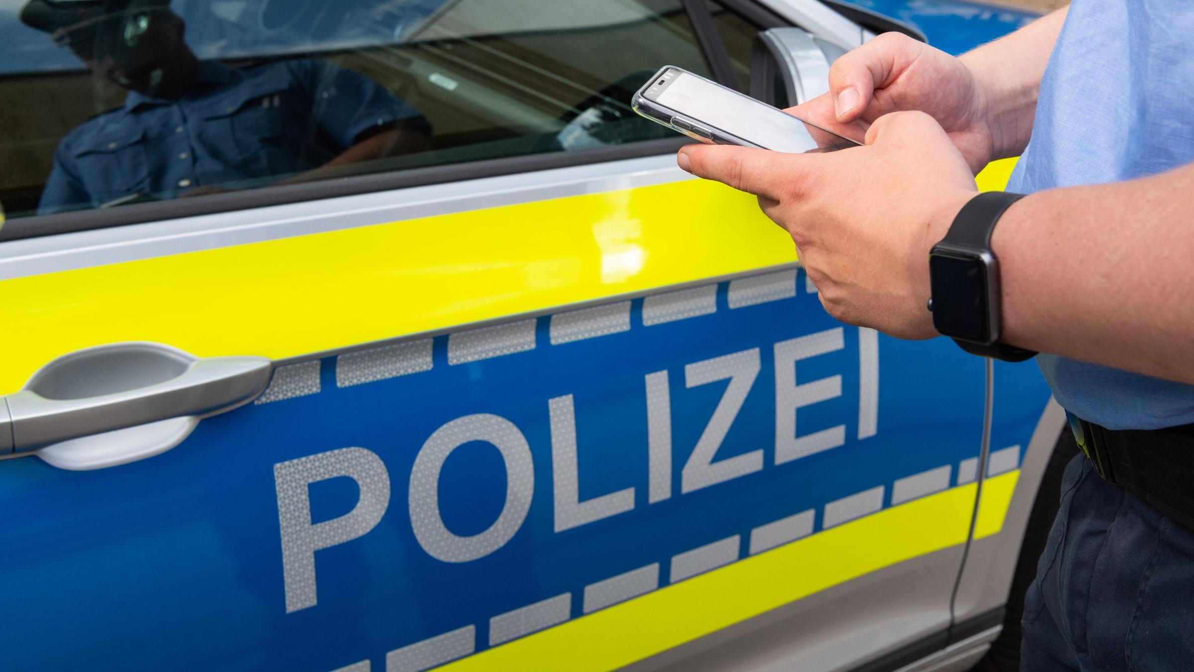 Ein Polizist hält sein Smartphone vor einem Polizeiauto. Symbol