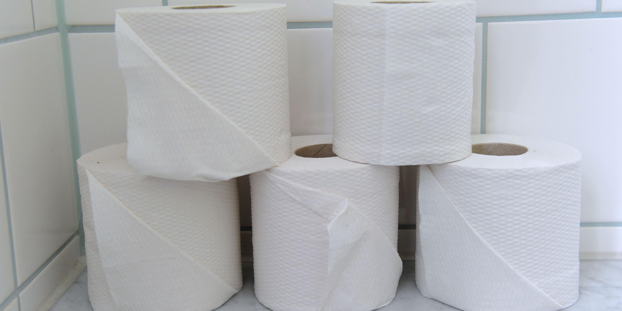 Auf einer gefliesten Ablage in einer Toilette stehen fünf Rollen Klopapier. Das Toilettenpapier ist in zwei Ebenen gestapelt: unten drei Rollen, darauf stehen zwei weitere Klopapierrollen versetzt.