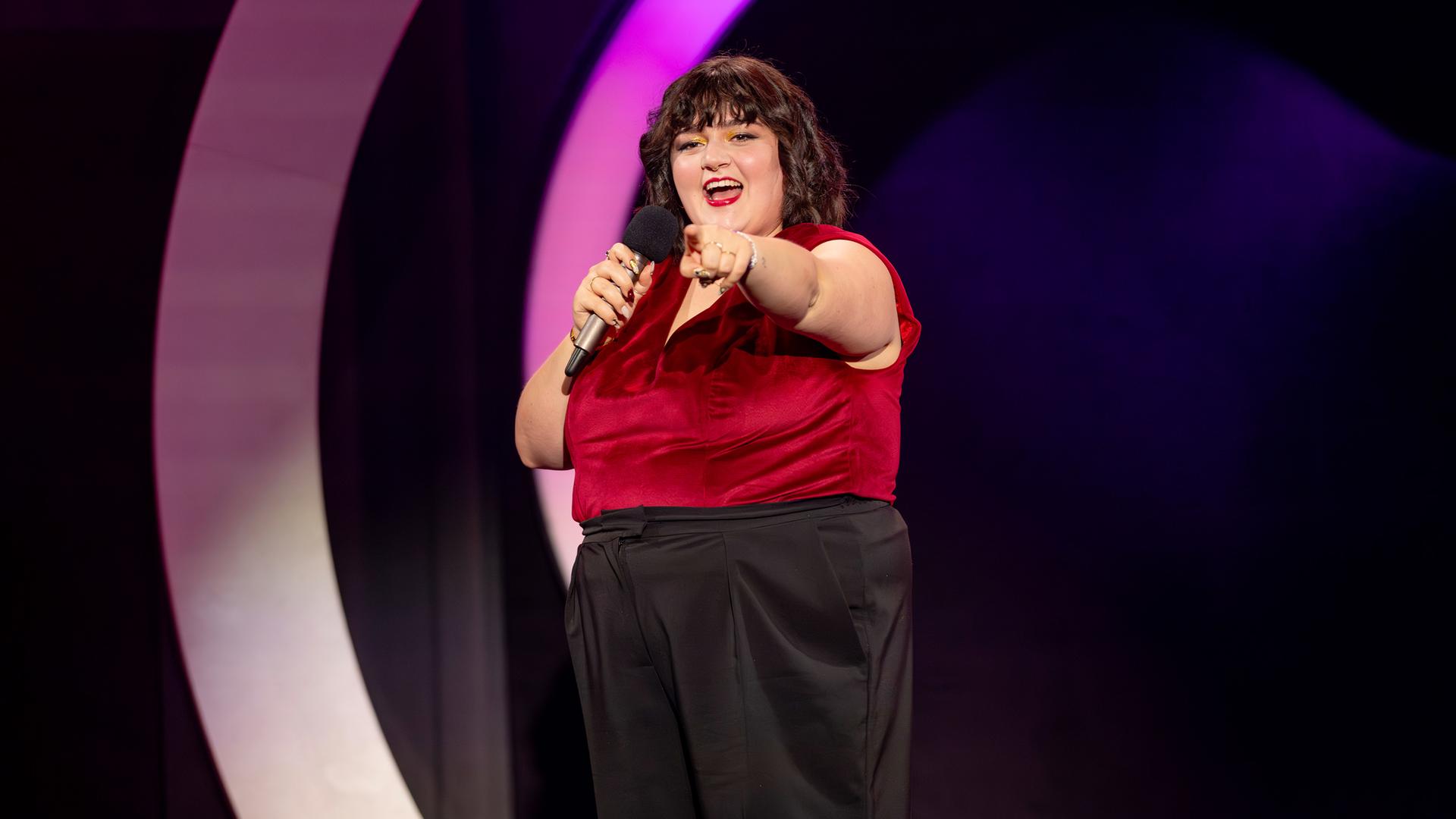 Teresa Reichl steht in der Mitte der Bühne. Sie streckt ihren linken Arm nach vorne aus. In der rechten Hand hält sie ein Mikrofon. Ihr Oberteil ist rot und sie trägt eine schwarze Hose.