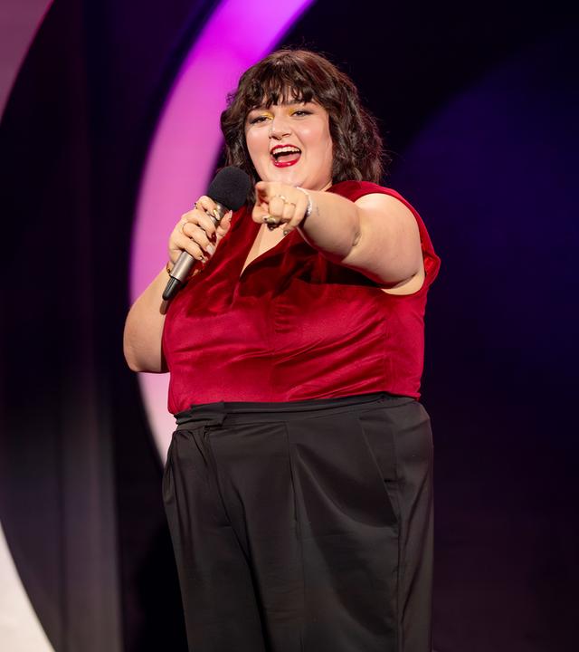 Teresa Reichl steht in der Mitte der Bühne. Sie streckt ihren linken Arm nach vorne aus. In der rechten Hand hält sie ein Mikrofon. Ihr Oberteil ist rot und sie trägt eine schwarze Hose.