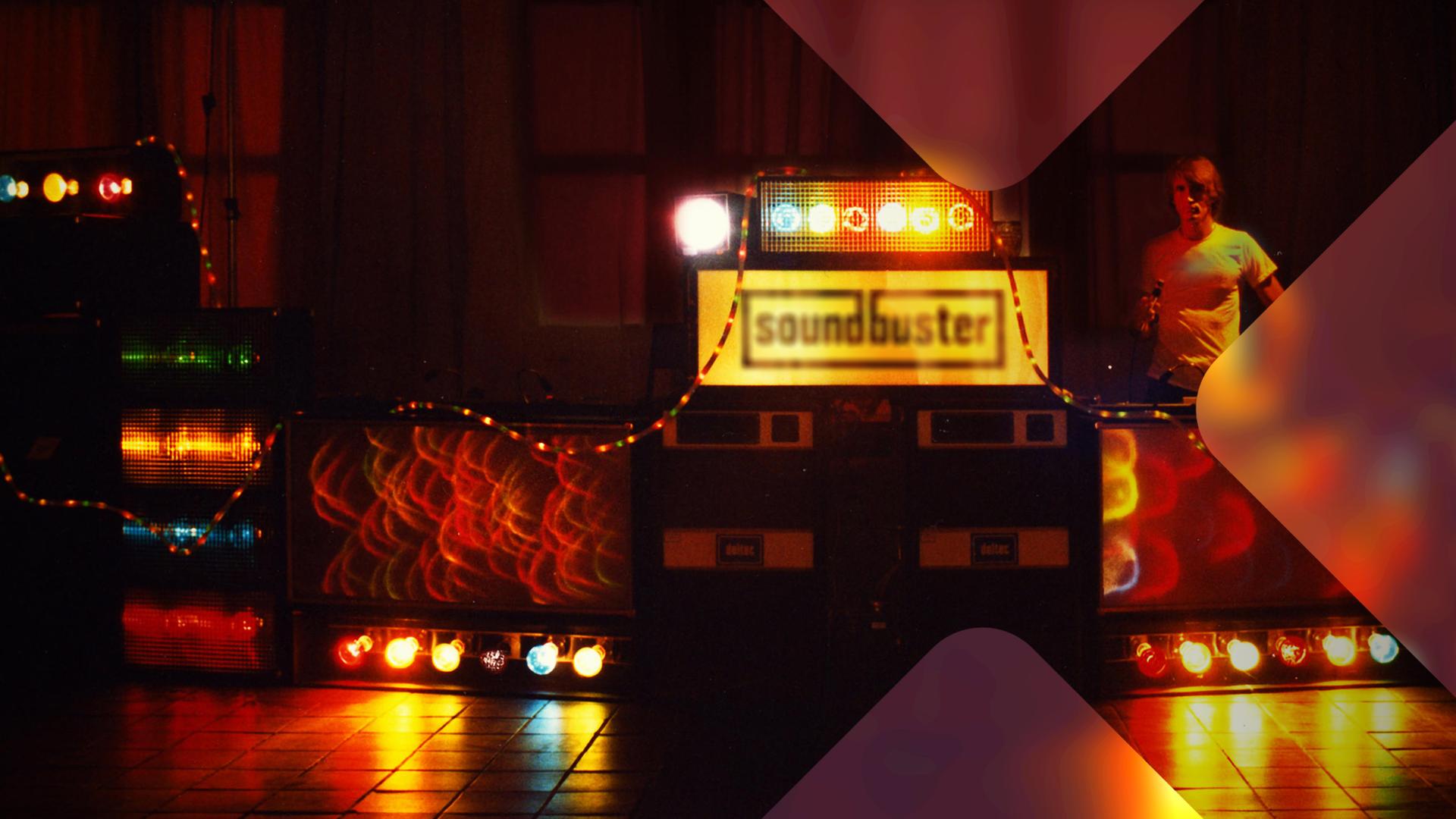 Ein Mann steht in einer dunkel beleuchteten Disco der 70er Jahre hinter einem Soundsystem. 