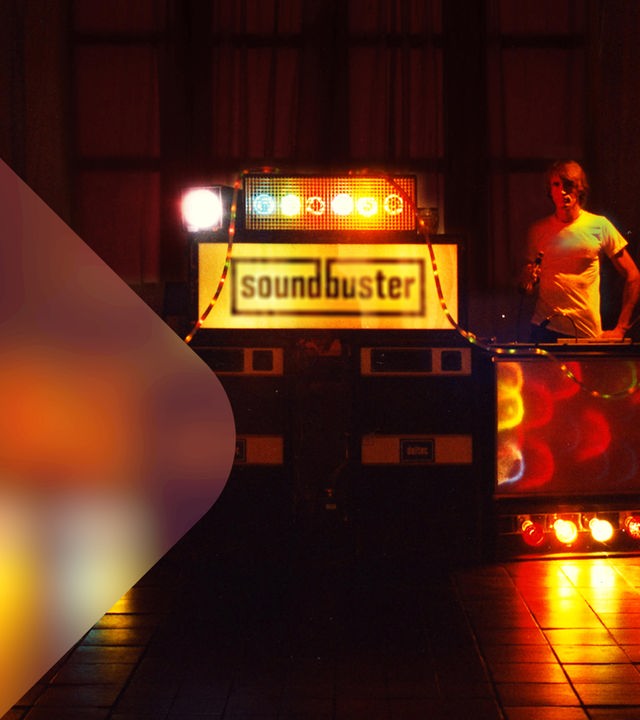 Ein Mann steht in einer dunkel beleuchteten Disco der 70er Jahre hinter einem Soundsystem. 
