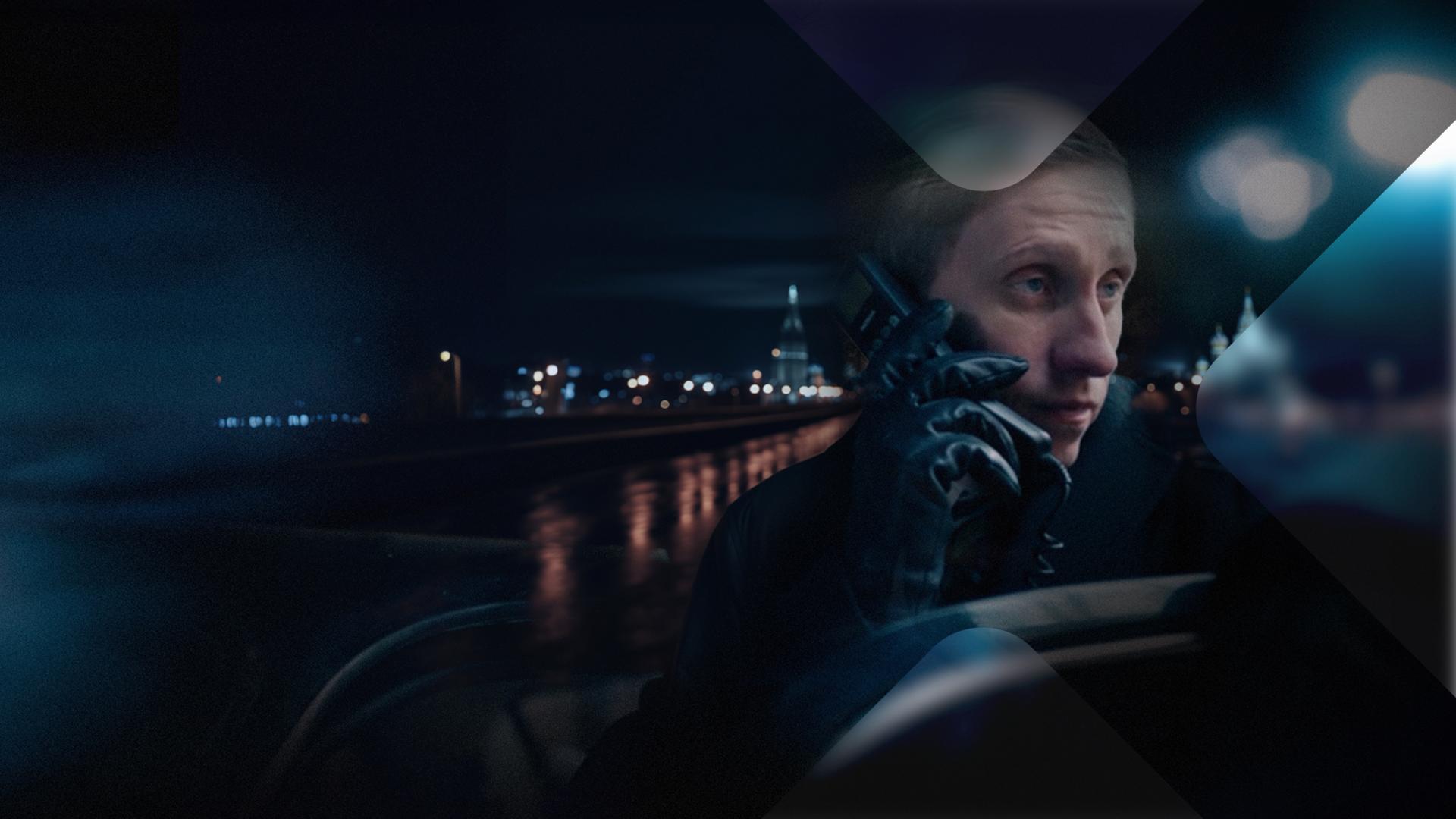 Putin Schauspieler im Profil mit Handy und Lederhandschuh vor dunklem Hintergrund.