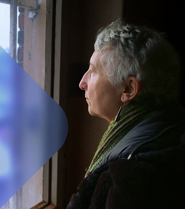 Eine Frau mit kurzen Haaren steht am Fenster eines Gefängnisses und blickt durch das vergitterte Fenster auf den Innenhof, in dem Schnee liegt. 