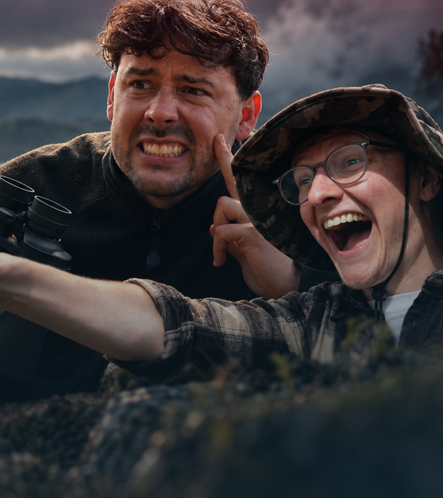Links im Bild erkennt man eine Trophäe mit einer kleinen Flagge auf einem Sockel. Daneben sind CrispyRob und CastCrafter vor einer dunklen, bergigen Landschaft montiert. CrispyRob hält ein Fernglas vor sich und verzieht angespannt das Gesicht. CastCrafter streckt den Arm zur Trophäe aus und schaut freudig.