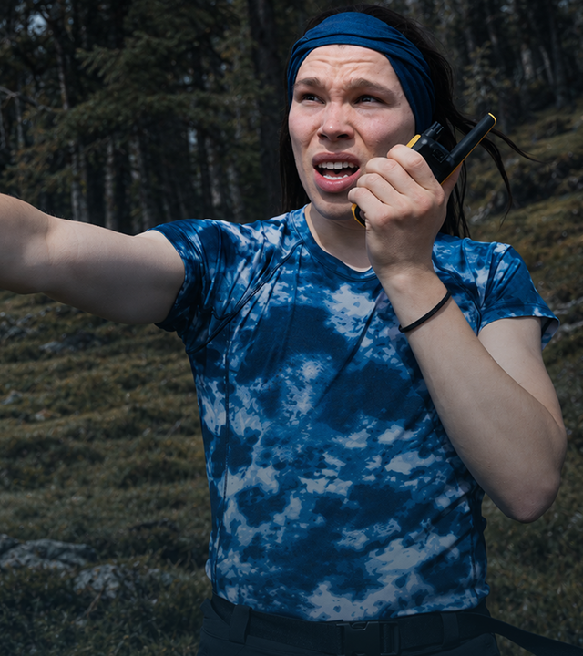 Lézan und Helge Mark stehen auf einem bewaldeten Hügel unter dunklen Wolken. Helge Mark befindet sich rechts im Vordergrund, trägt ein blau T-Shirt und ein blaues Stirnband. Er spricht in ein Funkgerät und zeigt mit ausgestrecktem Arm nach links. Weiter hinten steht Lézan mit einem roten Stirnband. Er hat beide Hände hinter dem Kopf verschränkt.