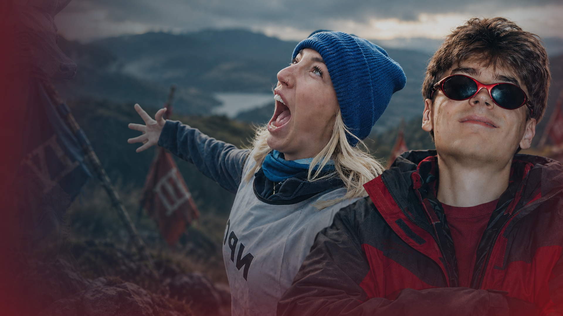 Starletnova steht links von Wichtiger in einer Berglandschaft. Sie trägt eine blaue Strickmütze, hat einen Arm ausgestreckt und blickt mit weit geöffnetem Mund nach oben. Wichtiger trägt eine rot-schwarze Jacke und eine Sonnenbrille.